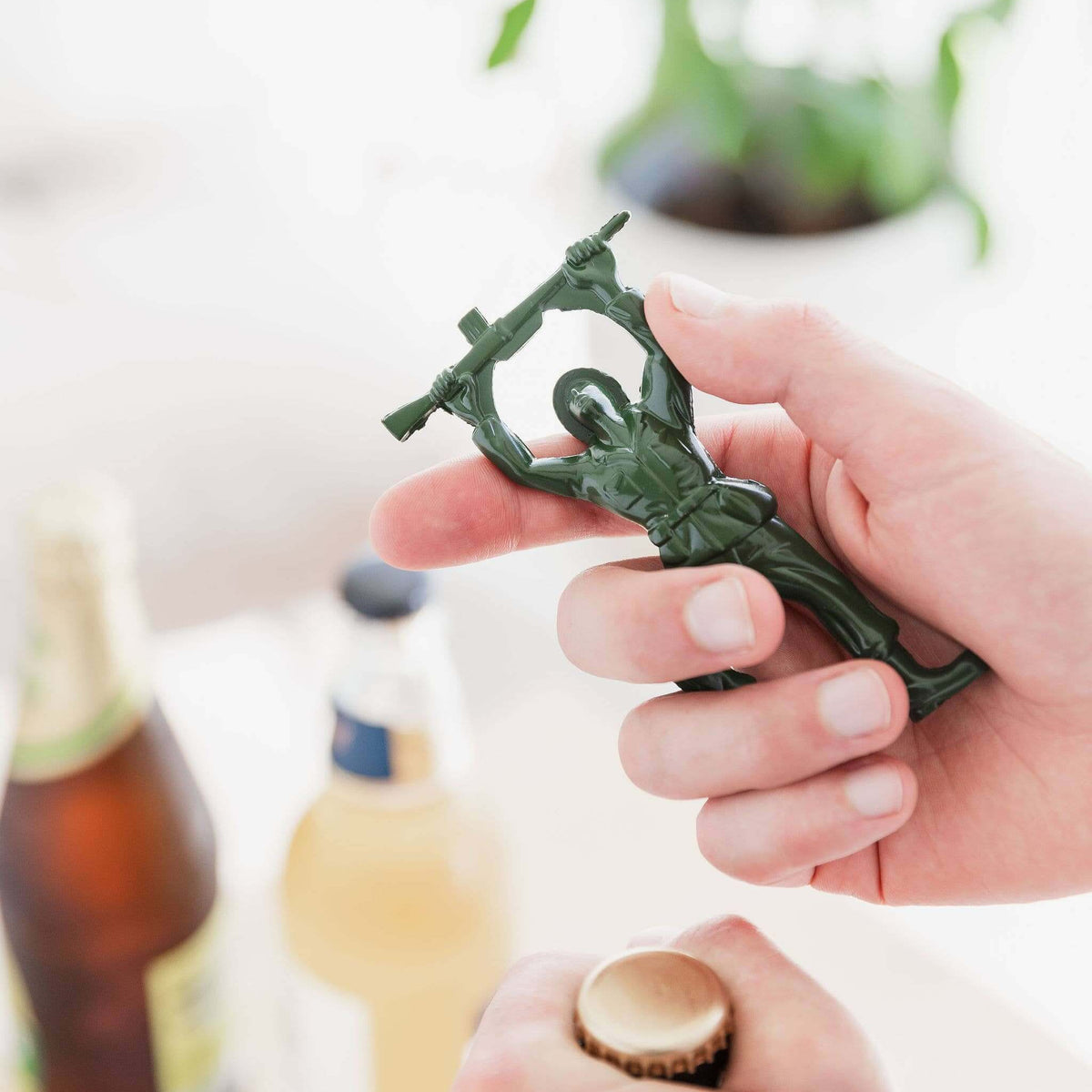 Hand holding a green bottle opener shaped like a person, with blurred bottles and plants in the background.