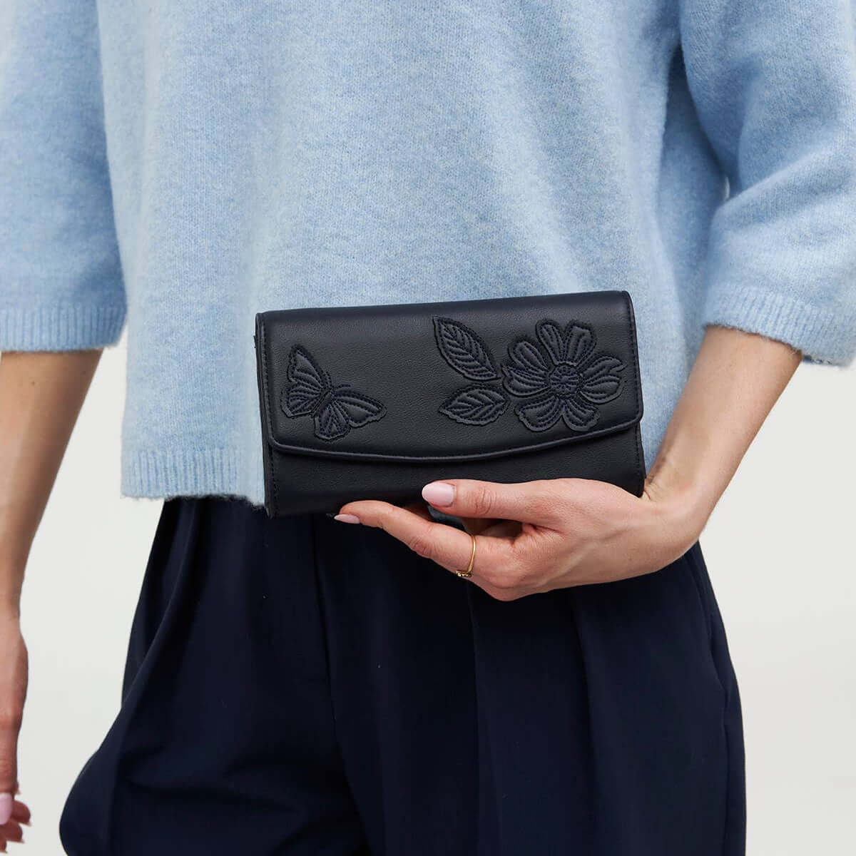 A person wearing a light blue knit sweater and dark navy trousers holds a black leather clutch bag with embossed floral and butterfly details on the front flap.