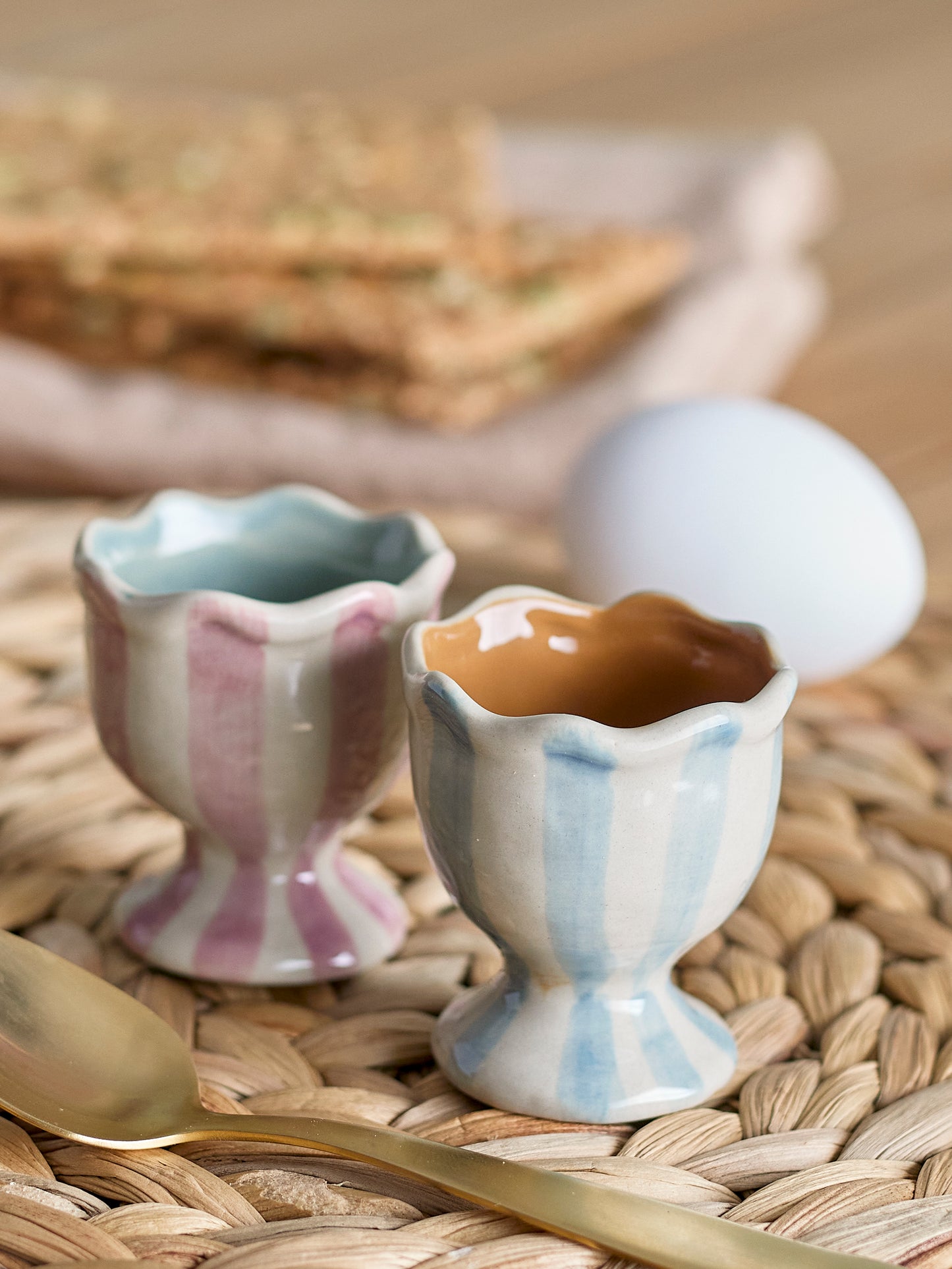 Two ceramic egg cups with a spoon and an egg on a woven mat