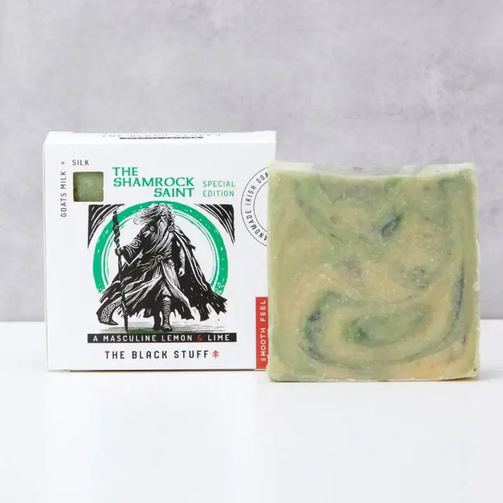 Packaging of 'The Shamrock Saint' natural soap by The Black Stuff featuring an illustration of a hooded figure holding a staff, with green accents and text indicating the product name and description. Un packaged bar of soap sitting alongside