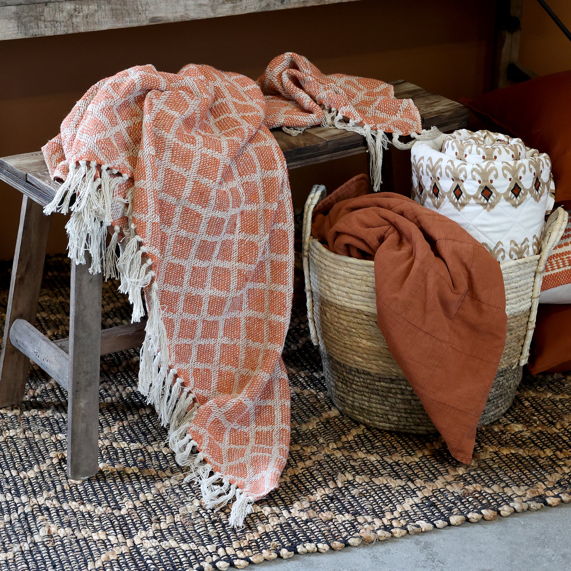 Decorative scene with a pink patterned blanket draped over a wooden stool and a woven basket with a brown blanket inside, on a textured rug.