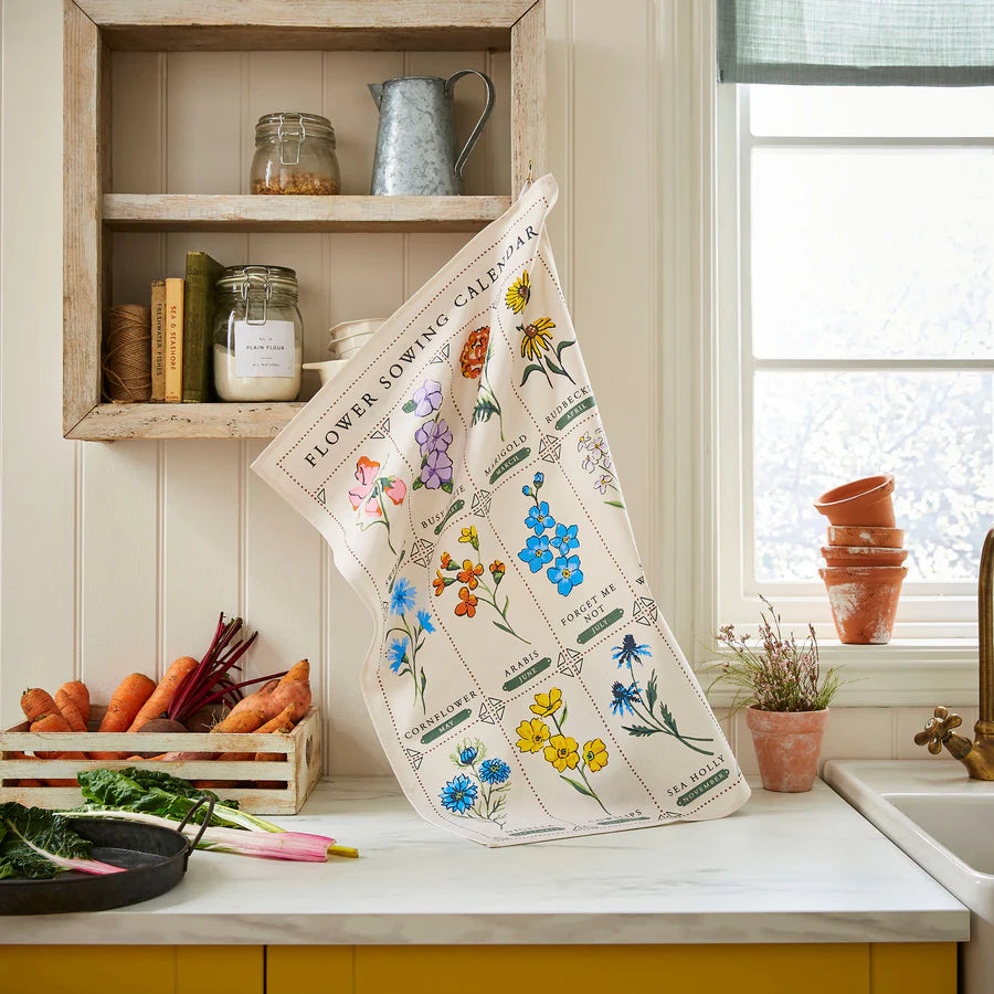 Floral-themed kitchen towel on a kitchen counter with a window and shelves in the background.