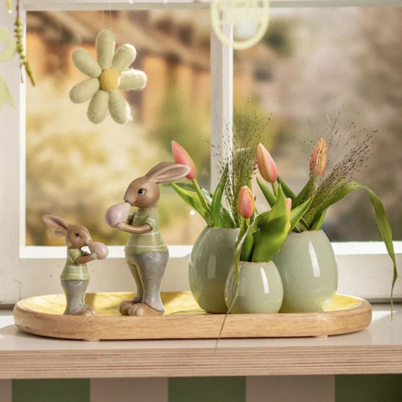 Decorative setup with rabbit figurines and tulips on a wooden tray in front of a window.
