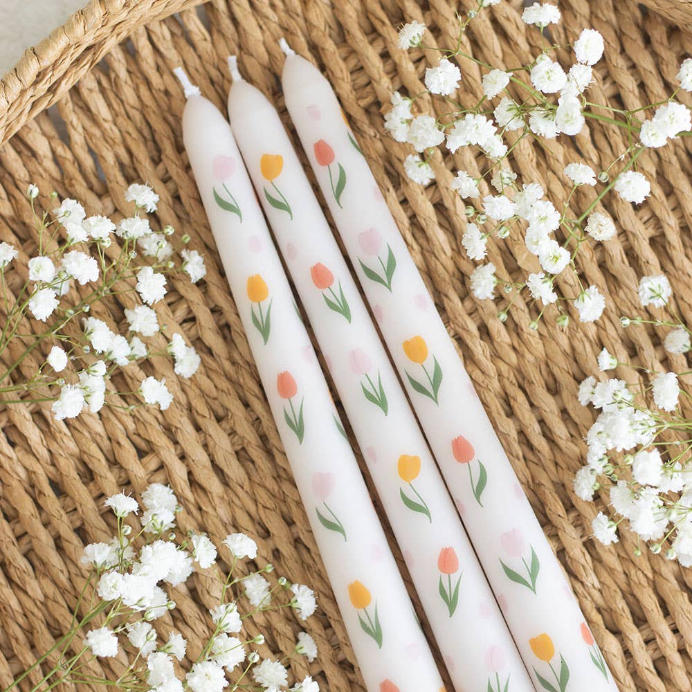 Three white candles with floral patterns on a woven surface with small white flowers.