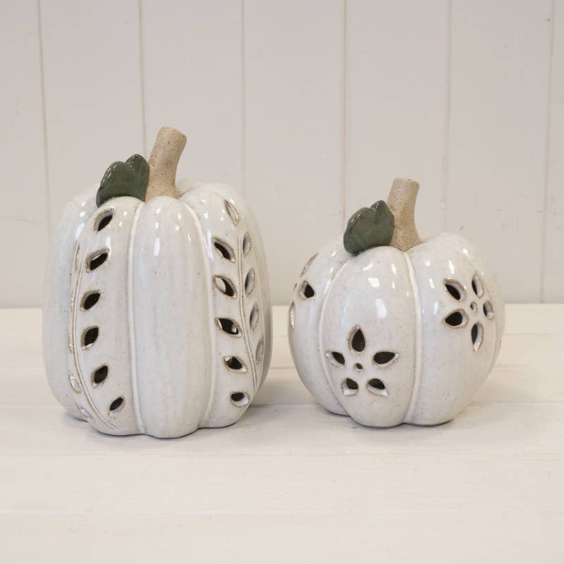 Two white ceramic decorative pumpkins with carved leaf and floral cutouts, featuring light brown stems and small green leaves, displayed on a white surface against a white paneled background.