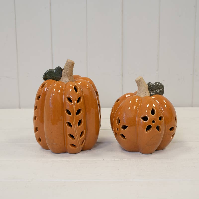 Two orange ceramic pumpkins with leaf-shaped cutouts and dark green leaves, displayed on a white surface against a white paneled background.