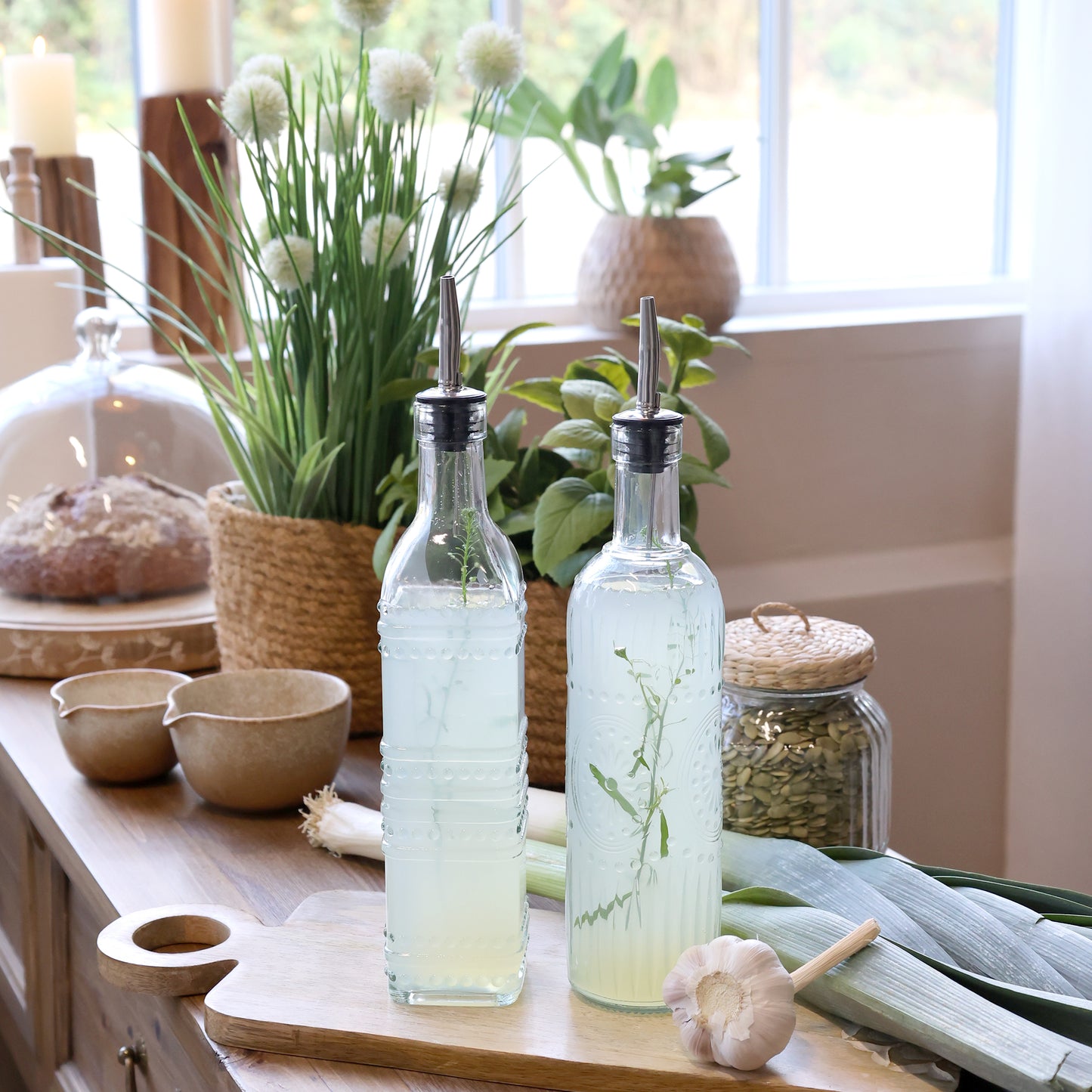 Two glass bottles with metal nozzles on a wooden surface with plants and kitchen items in the background.