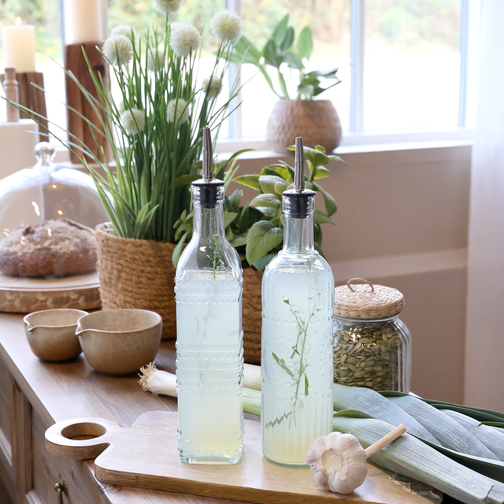 Two glass bottles with metal nozzles on a wooden surface with plants and kitchen items in the background.