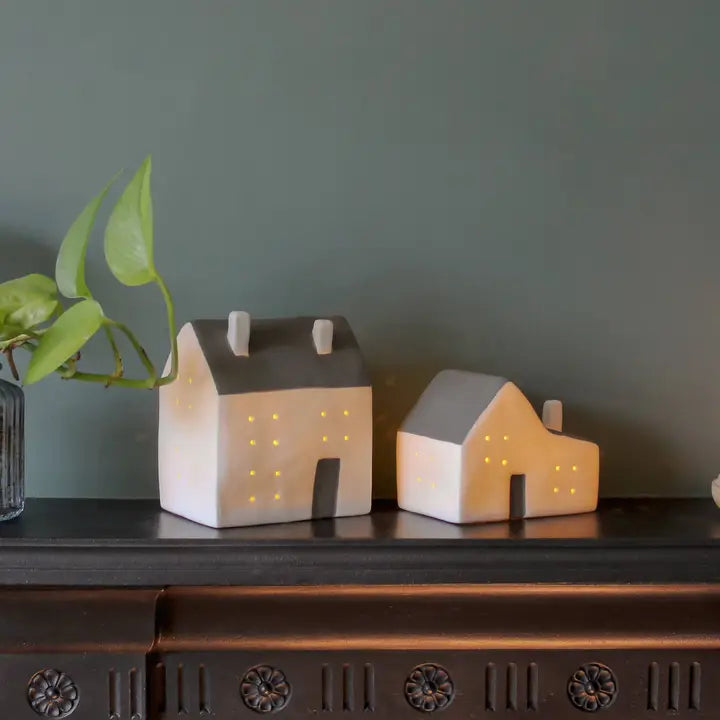 Two small house-shaped lamps on a dark surface with a green plant in the background.