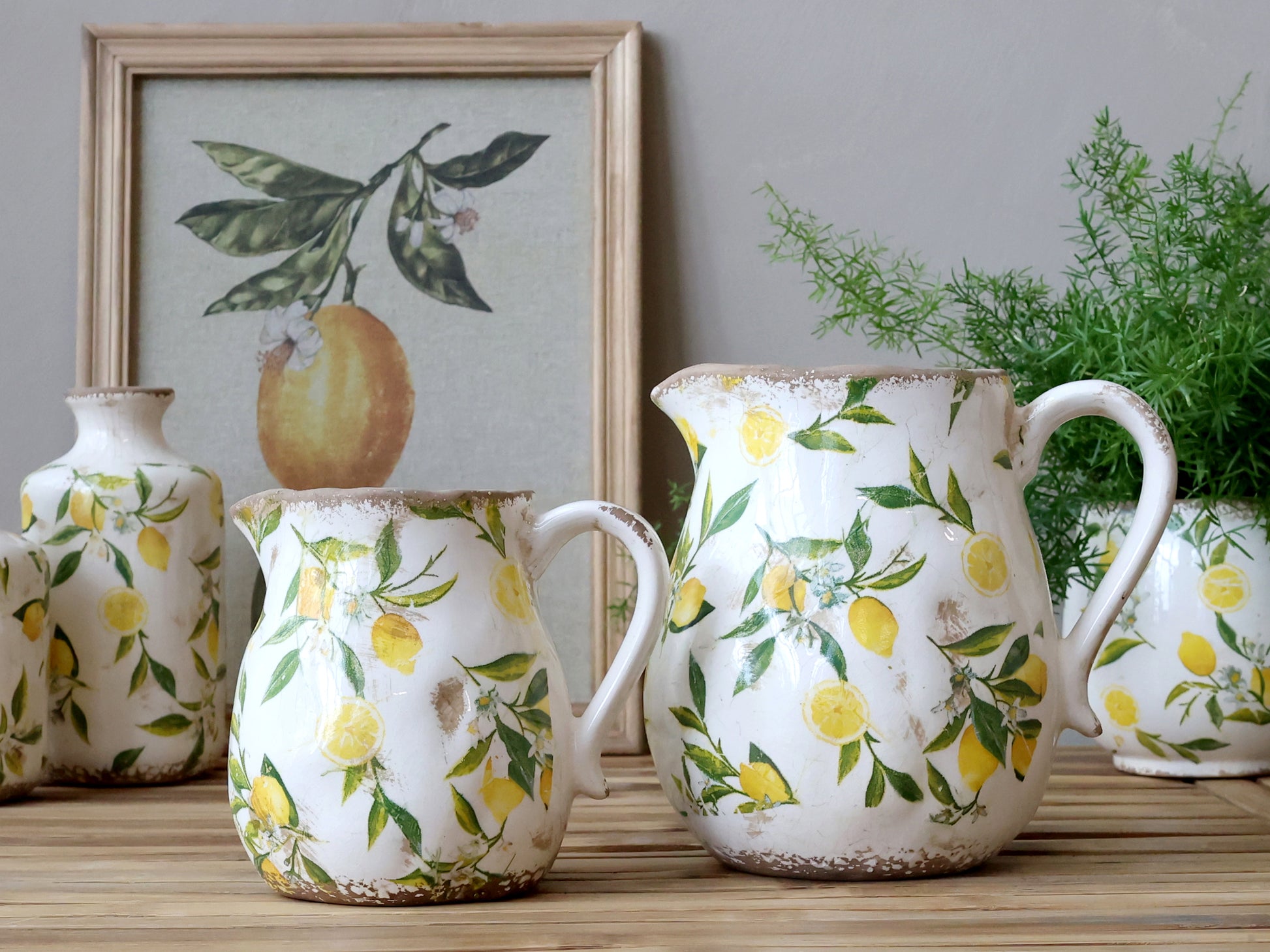 Set of ceramic jugs with lemon and leaf pattern on a wooden surface, framed artwork in the background.