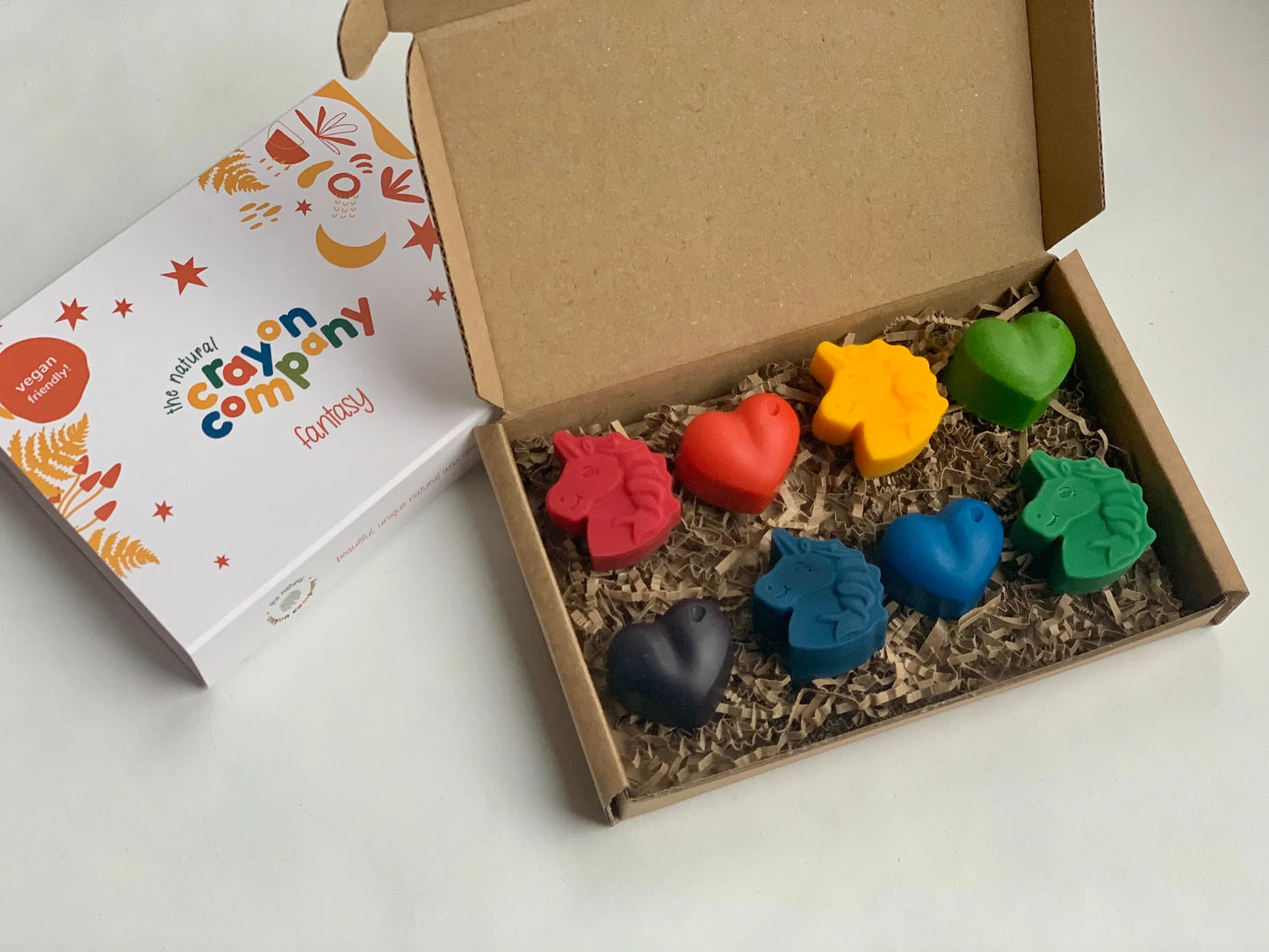 Colorful heart and unicorn shaped crayons in a box with a branded card on a white background