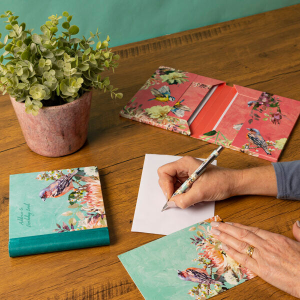 Person writing on a piece of paper with floral notebooks and a plant on a wooden table.