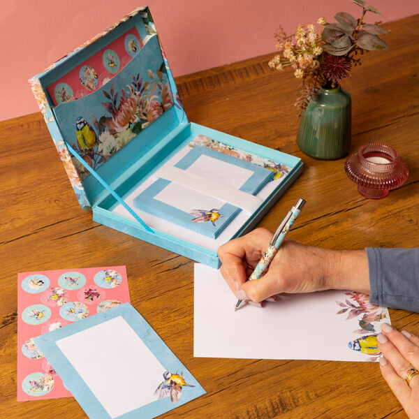 Person writing with a pen on a piece of paper next to a decorative stationery set on a wooden table.
