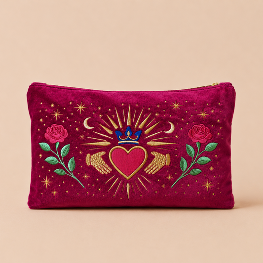 A magenta velvet zippered pouch featuring a detailed embroidered design of a central red heart with a blue crown, gold rays, wings, and stars, flanked by pink roses and green leaves, set against a solid beige background.