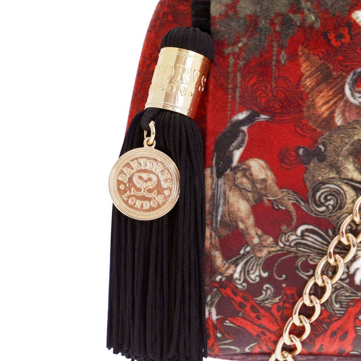 Close-up of a luxury handbag featuring a prominent black tassel with a gold-toned 'FANTASY LONDON' medallion charm, set against a red and brown patterned fabric background with a gold chain strap.