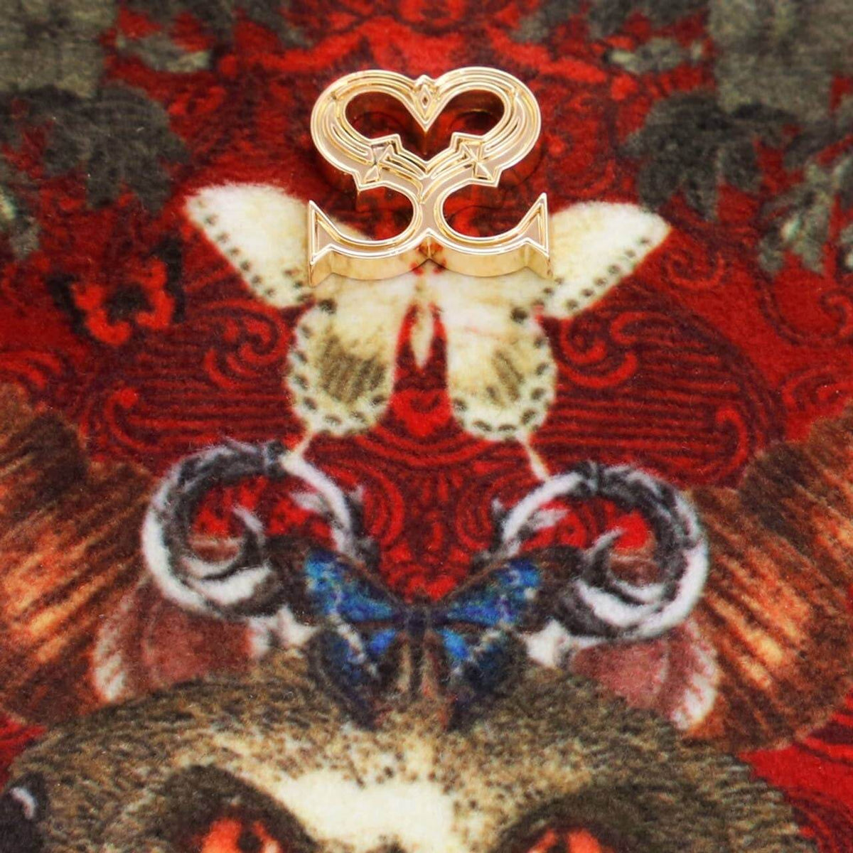 A gold-toned double-heart ring with a stylized interlocking 'SC' design, displayed on a highly detailed red and brown brocade fabric background.
