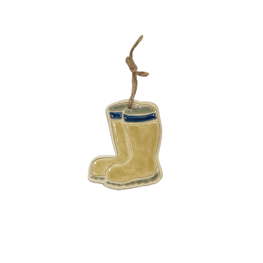 A ceramic decoration in the shape of a pair of mustard colored wellies with navy trim, hanging from a jute string.