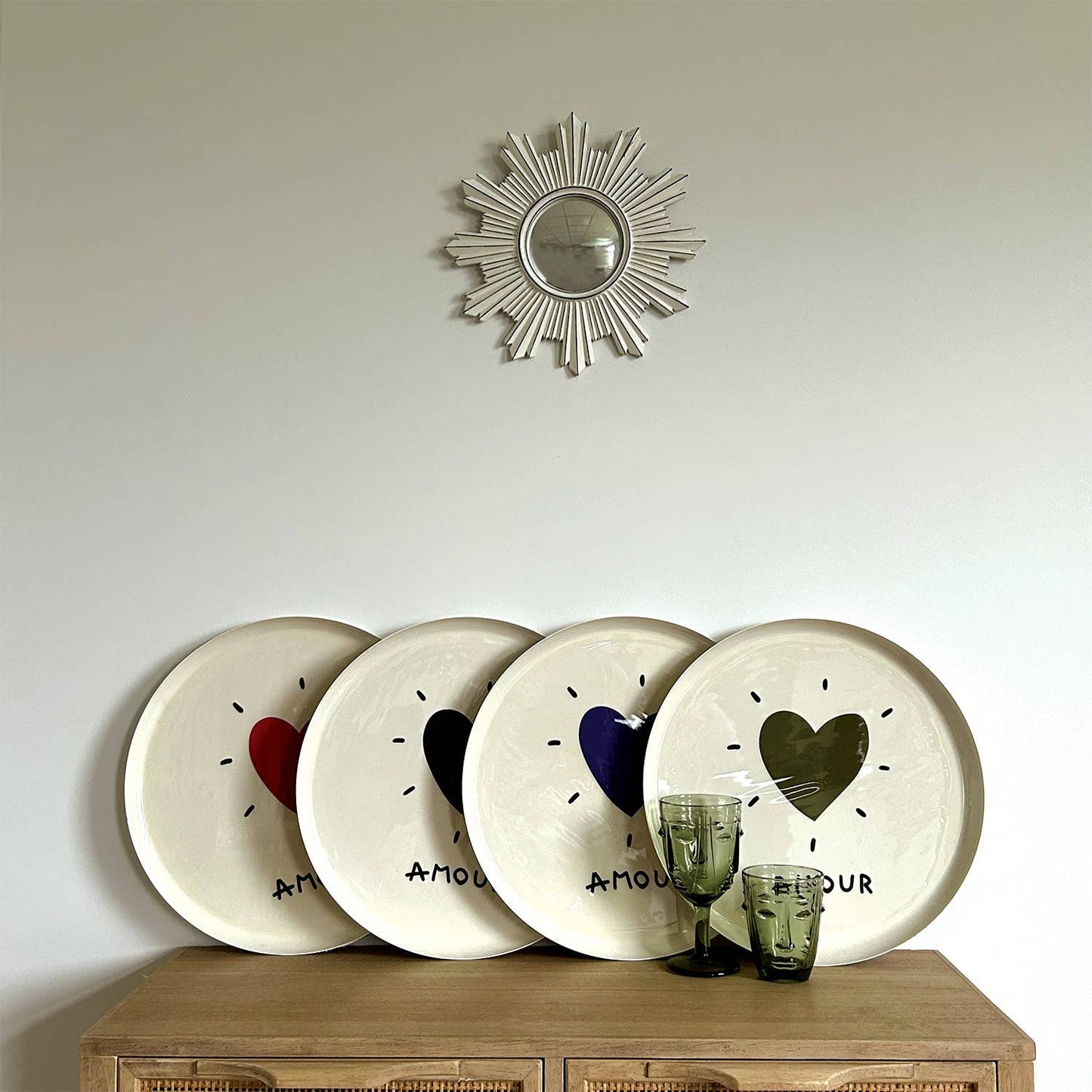 Four decorative plates with heart designs on a wooden cabinet against a light wall with a sunburst mirror.