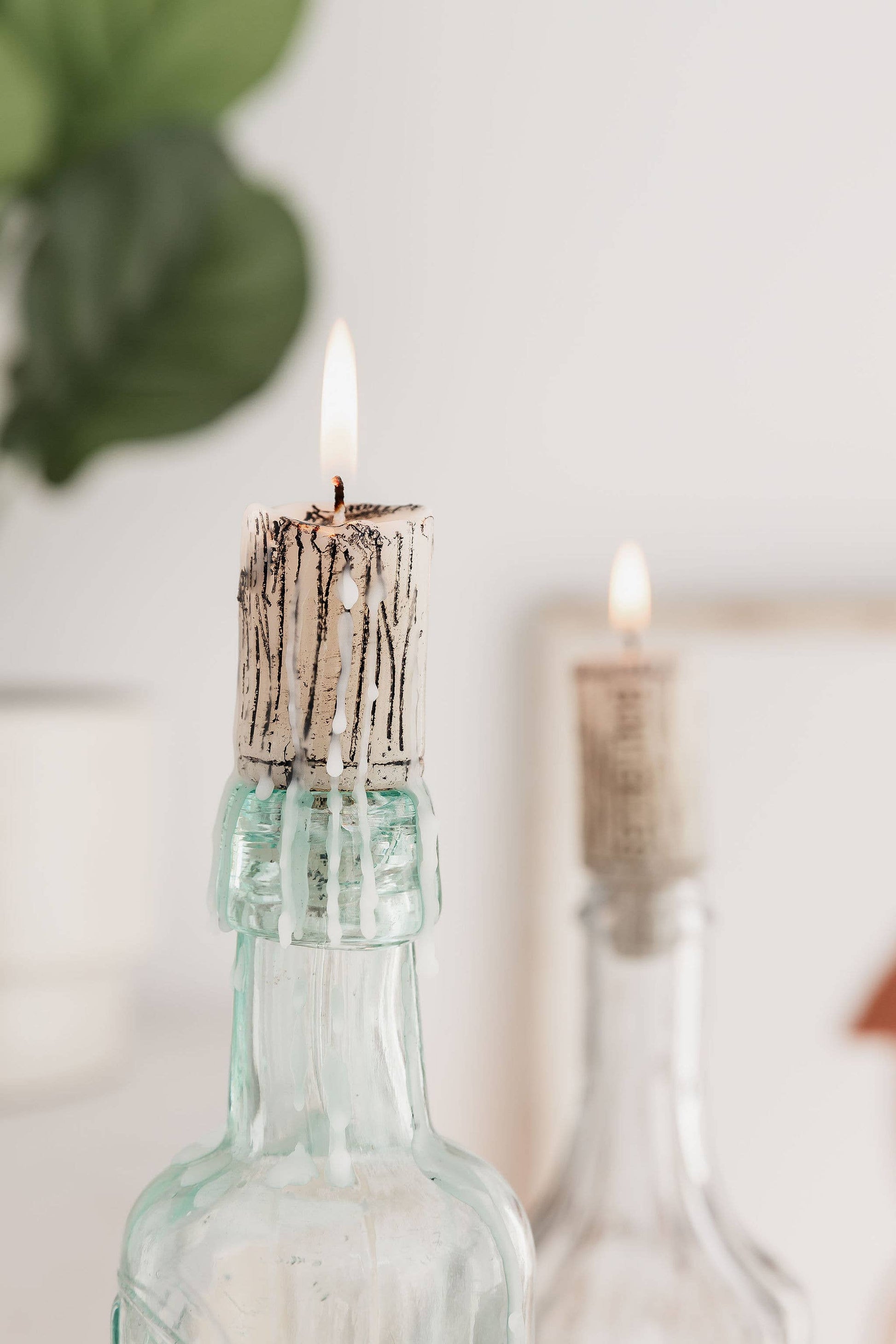 Candle in a glass bottle with a textured wick, surrounded by blurred greenery.