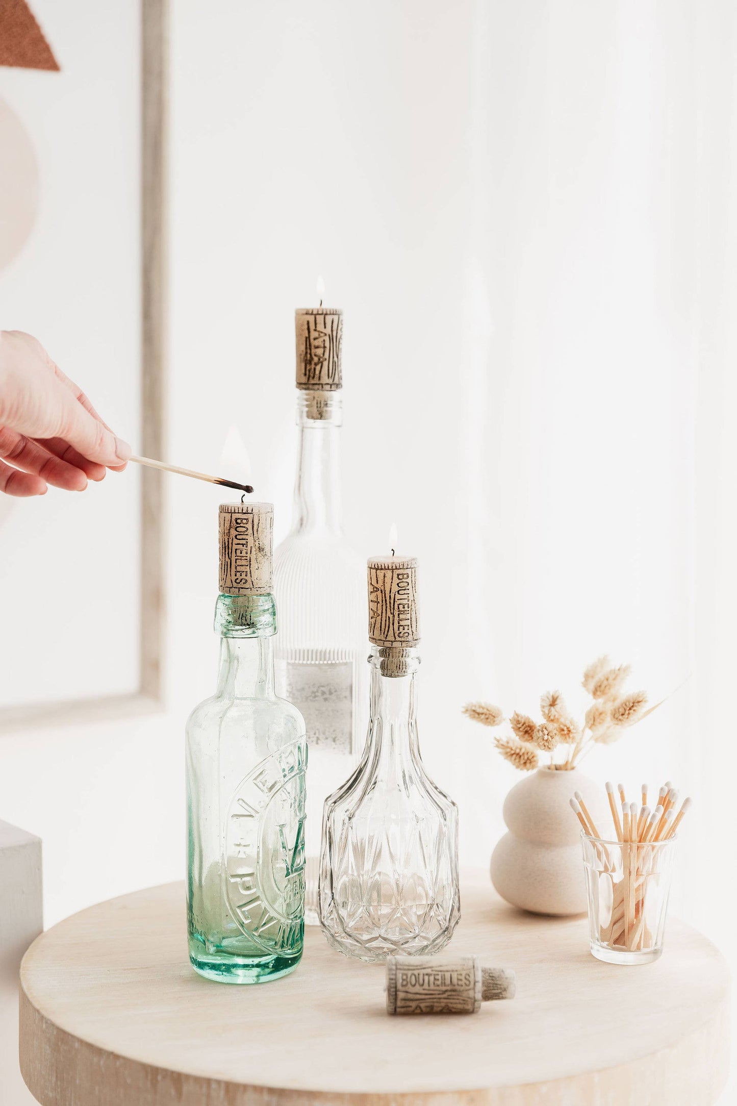 Two glass bottles with cork candles on a wooden surface, with a hand holding a match.
