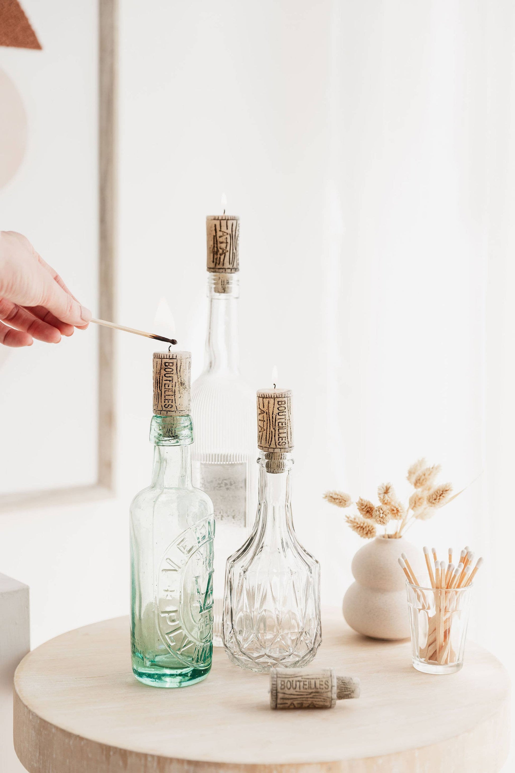Two glass bottles with cork candles on a wooden surface, with a hand holding a match.