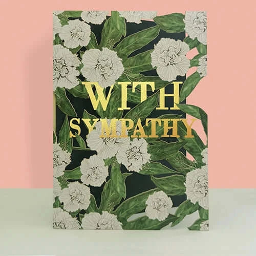 A rectangular greeting card with a floral pattern of white flowers and green leaves, featuring the words 'WITH SYMPATHY' in large gold letters, set against a split pink and white background.