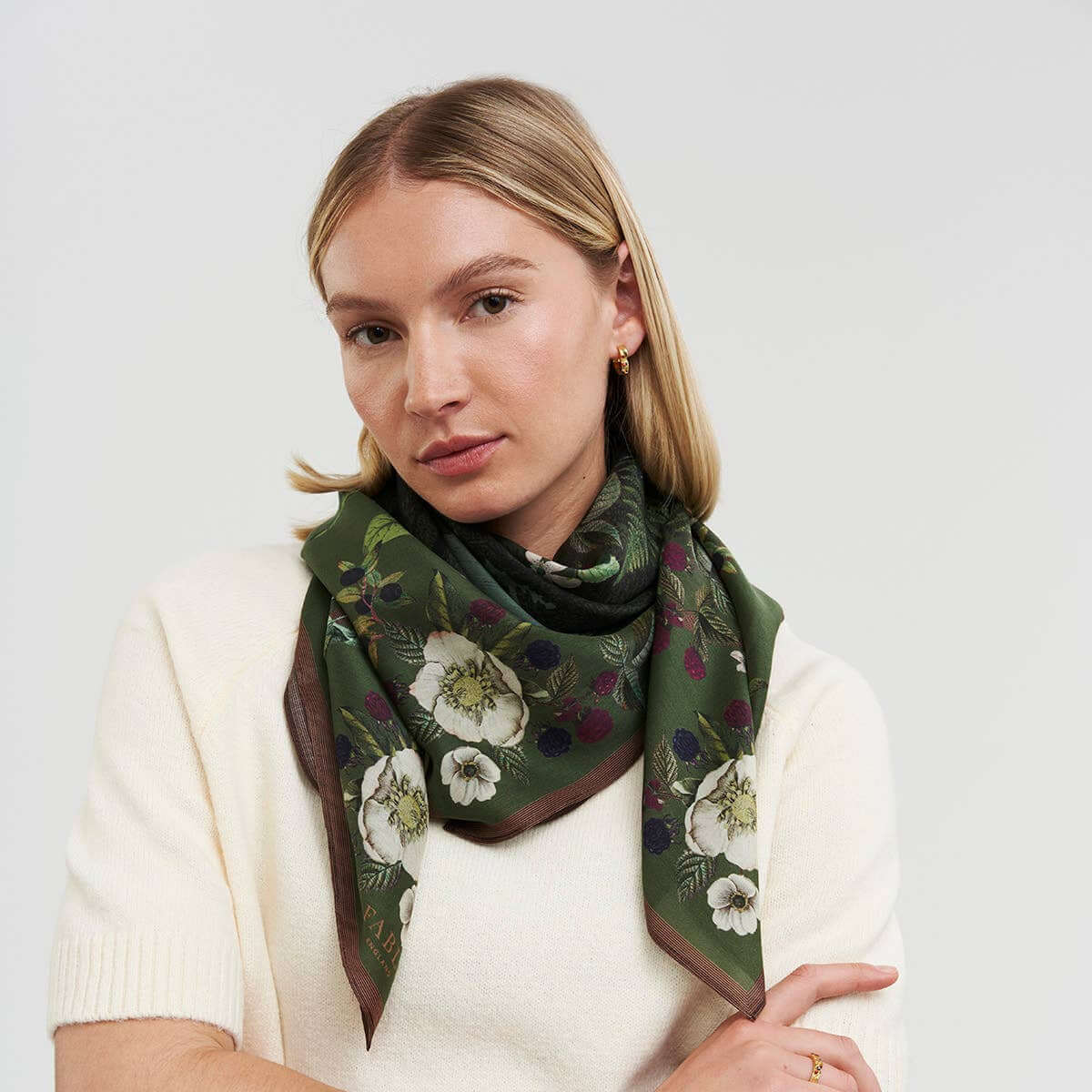 A woman wearing a cream-colored knit sweater and a dark green floral silk scarf with white and purple flowers, posed against a light gray studio background.