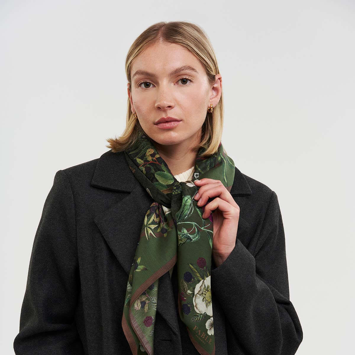 Woman wearing a dark coat with a green floral scarf around her neck against a white background