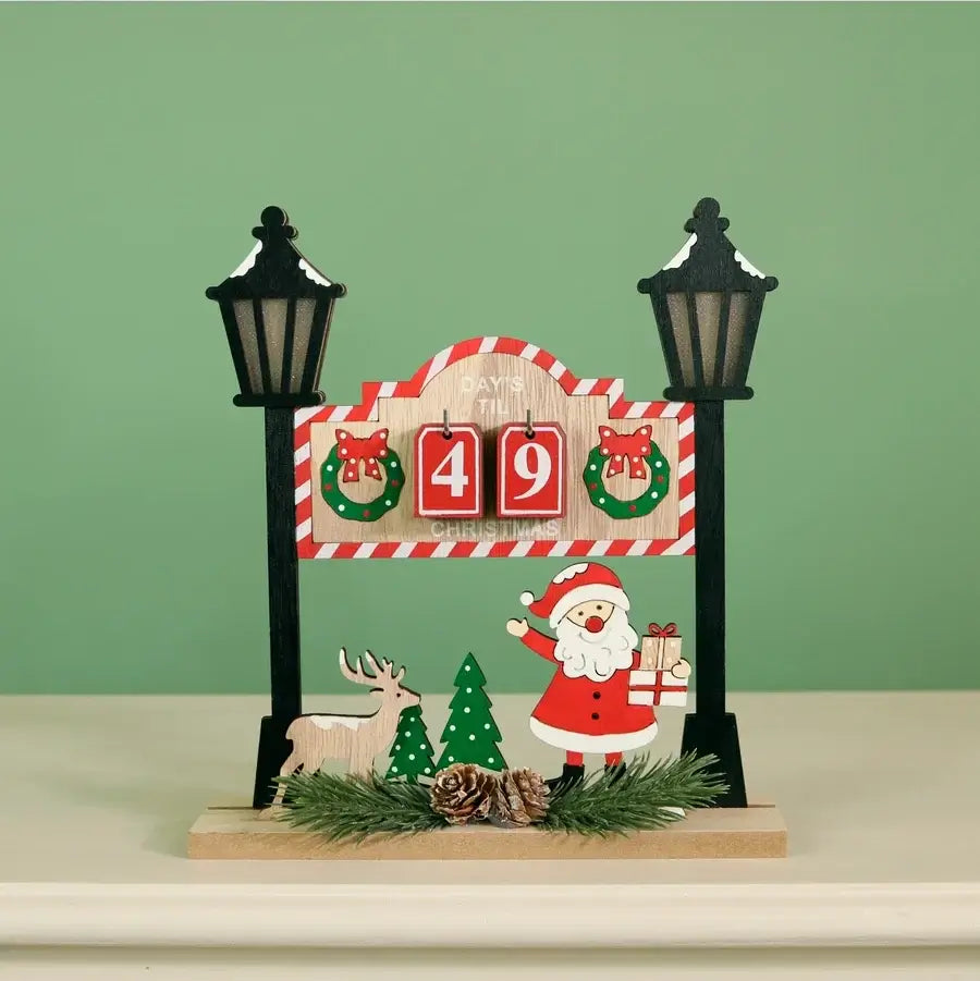 A Christmas-themed tabletop decoration featuring a wooden sign with red-and-white striped trim, two black lamp posts, a Santa Claus figure, a reindeer, pine trees, and a red '49' block display, set on a light surface against a pale green background.