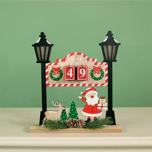 A Christmas-themed tabletop decoration featuring a wooden sign with red-and-white striped trim, two black lamp posts, a Santa Claus figure, a reindeer, pine trees, and a red '49' block display, set on a light surface against a pale green background.