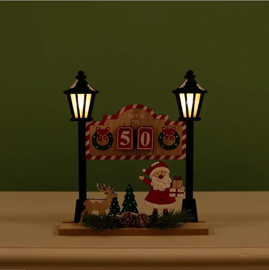 A decorative Christmas tabletop sign with two black lantern posts, a wooden board reading 'Days to the 50', a Santa Claus figure, a reindeer, pinecones, evergreen branches, and a small Christmas tree, set against a dark green background.