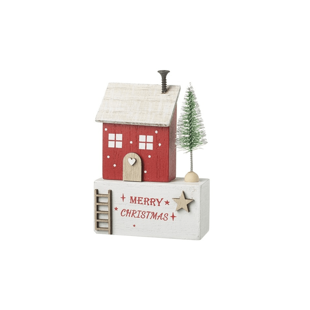 A small Christmas house figurine with a red body, beige roof, white base, and a small green tree beside it. The front of the base reads “MERRY CHRISTMAS” with a star detail, photographed on a plain white background.