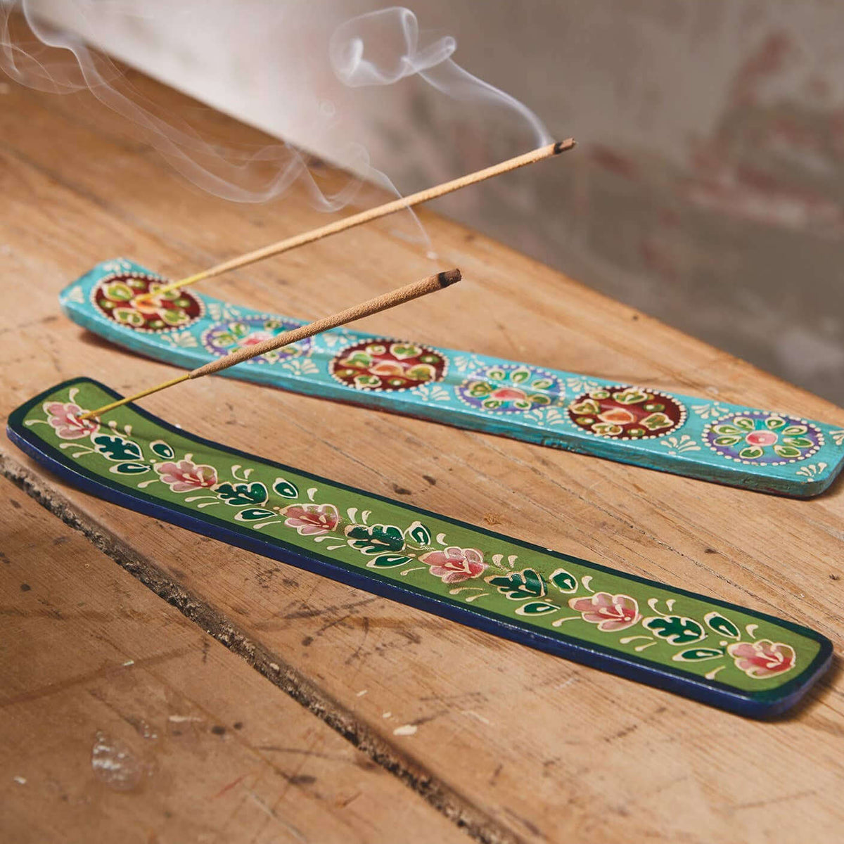 Two decorative incense burners with floral patterns, one turquoise and one green, holding incense sticks with visible smoke on a wooden surface.