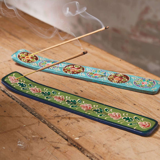 Two decorative incense burners with floral patterns, one turquoise and one green, holding incense sticks with visible smoke on a wooden surface.