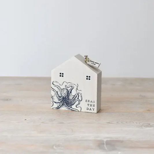 A wooden house-shaped ornament with a nautical theme, featuring an octopus and the phrase 'Seas the Day' printed on it.