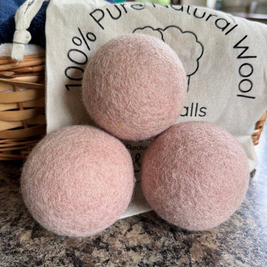 Three pink wool dryer balls on a countertop with a bag labeled '100% Pure Natural Wool Balls' in the background.