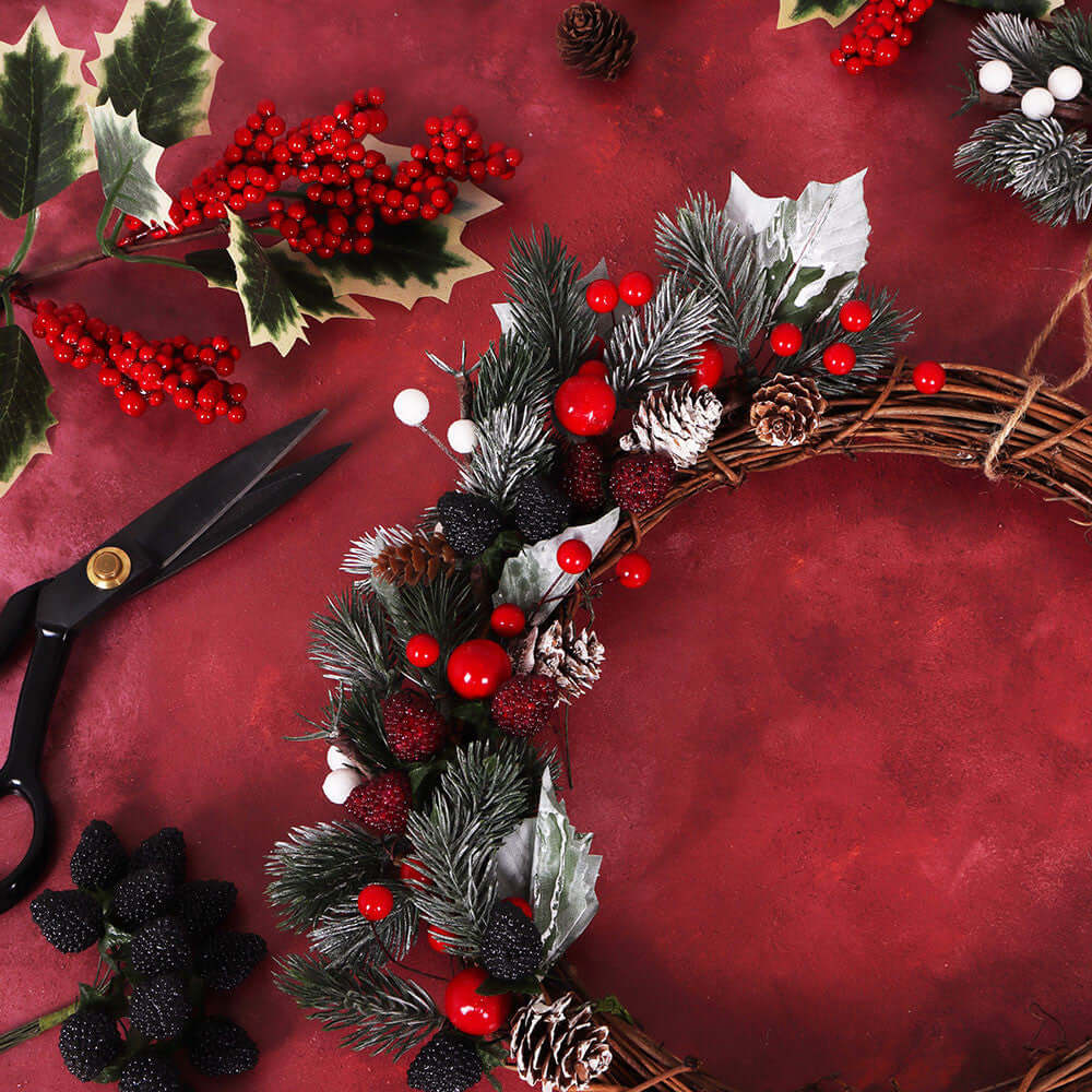 A festive Christmas wreath made of woven twig branches, decorated with artificial pine needles, red berries, white berries, pinecones, and holly leaves, arranged on a textured red background with scissors and holiday foliage nearby.