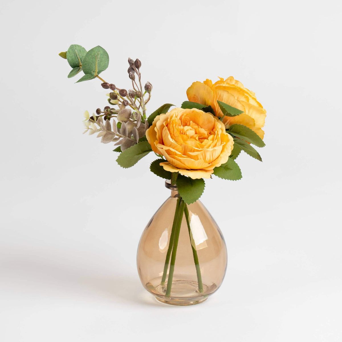 Yellow artificial roses and green foliage arranged in a small teardrop-shaped clear glass vase on a light gray background.