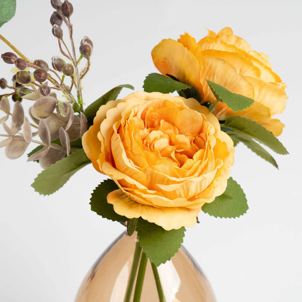 A close-up of an artificial floral arrangement featuring two large yellow-orange roses, green leaves, and small muted gray-green sprigs in a clear glass vase against a light gray background.