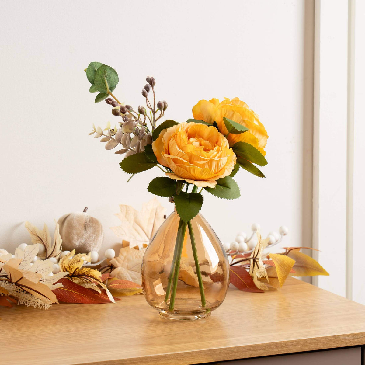 A clear glass vase holding a yellow-orange faux rose bouquet with green leaves, styled on a light wood table against a white wall with autumn decorative leaves around it.