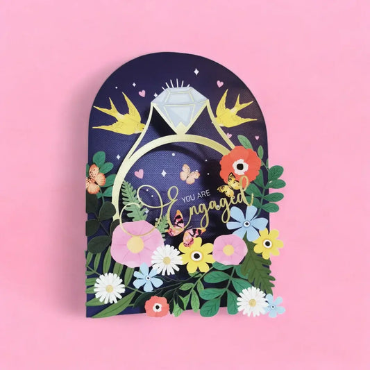 A 3D papercraft sign shaped like an arch with a navy blue background, featuring a gold ring with a light blue diamond, yellow birds, colorful flowers, butterflies, and the words 'You are Engaged' in gold script, set against a solid pink background.