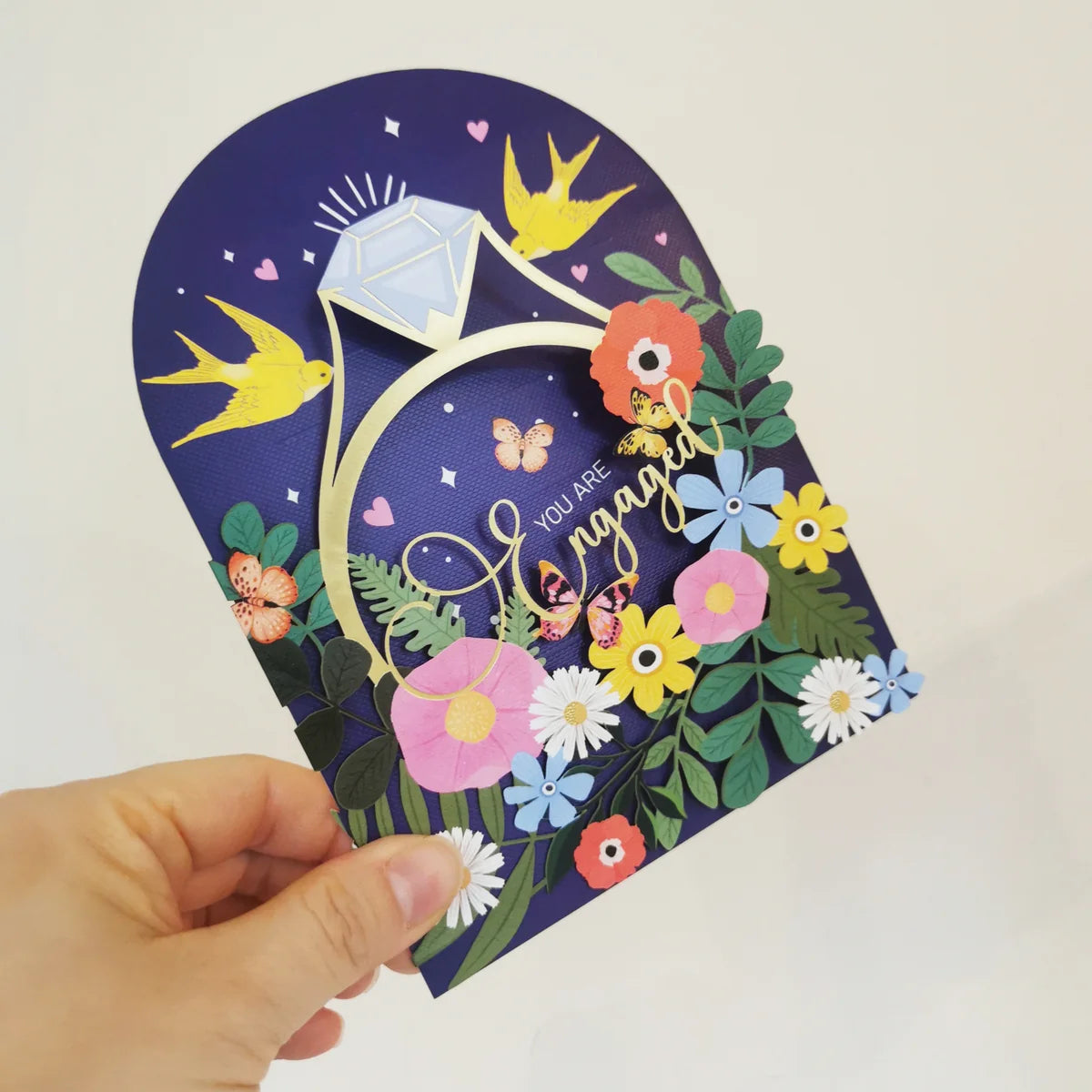 A hand holds a 3D layered pop-up engagement card featuring a large diamond ring, colorful paper flowers, butterflies, and yellow birds on a dark blue background.