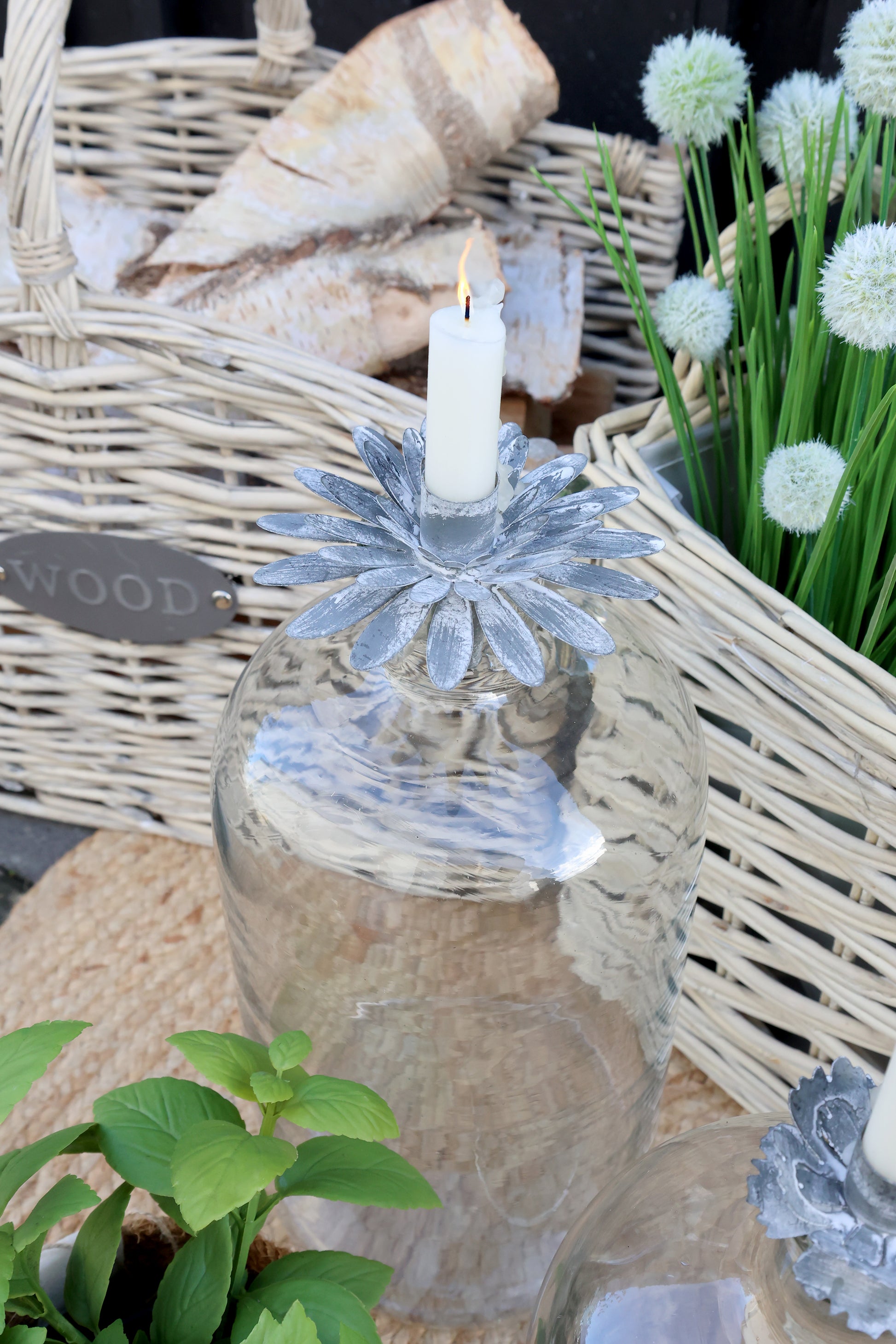 Decorative glass candle holder with a candle, surrounded by wicker basket and plants.