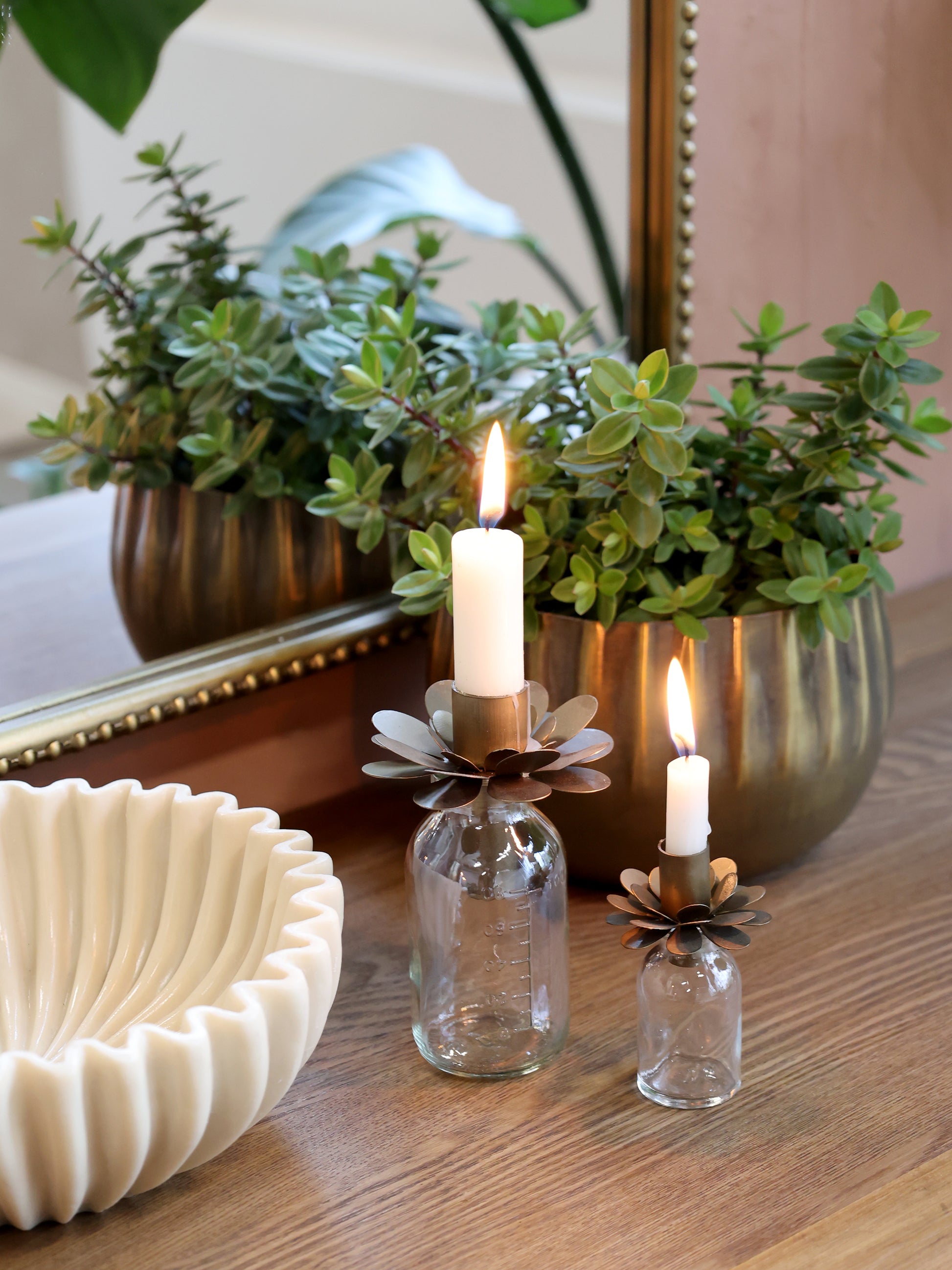 Two small glass candle holders with candles on a wooden surface, with plants and a mirror in the background.