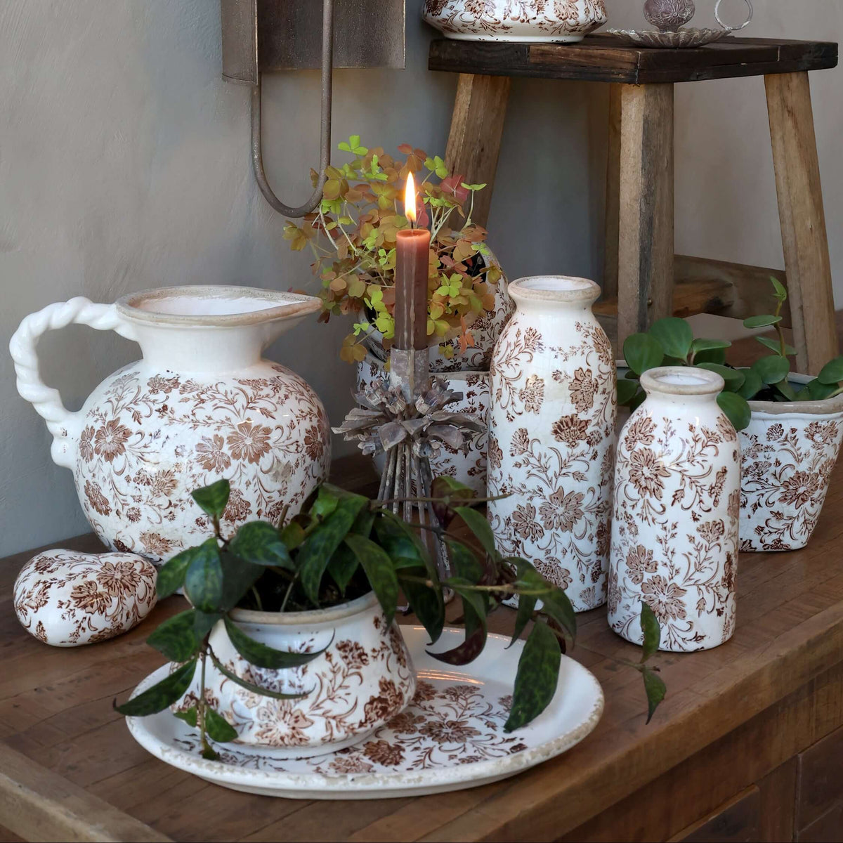 A styled tabletop arrangement of white ceramic vases and a pitcher with a brown floral toile pattern, set on a wooden table with lit candles and small potted plants against a textured gray wall.