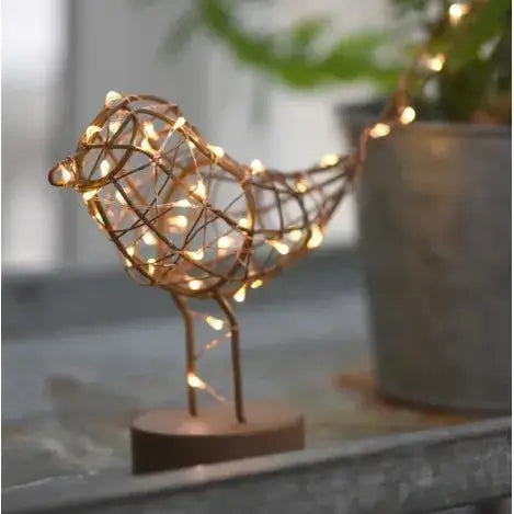 A decorative bird-shaped wire sculpture with warm white string lights, mounted on a round wooden base and photographed on a tabletop with a blurred indoor background and potted plants.